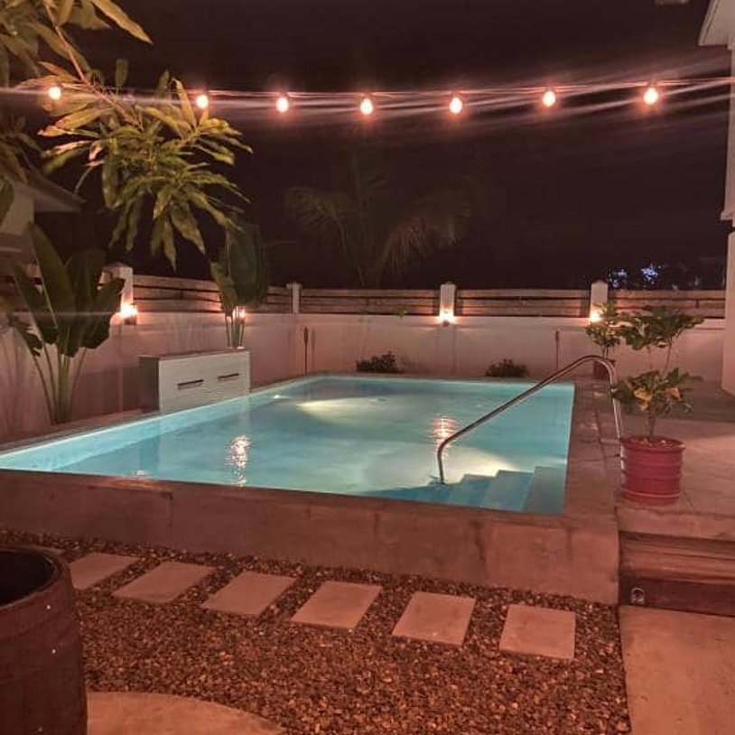 Night time, lights are lighting up the backyard, over the pool, bbq and outdoor chairs